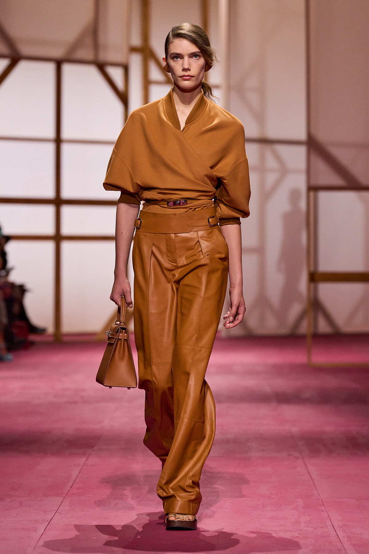 Model walks a runway in caramel-brown, oversized wrap-top with high-waisted leather pants and a small tan handbag.