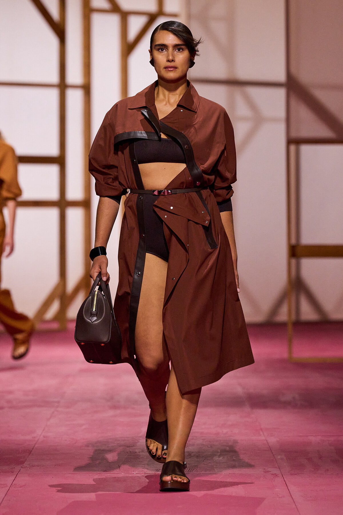Model walking a fashion show in a brown trench over a black crop top, with a cinching belt and a black handbag on her arm.
