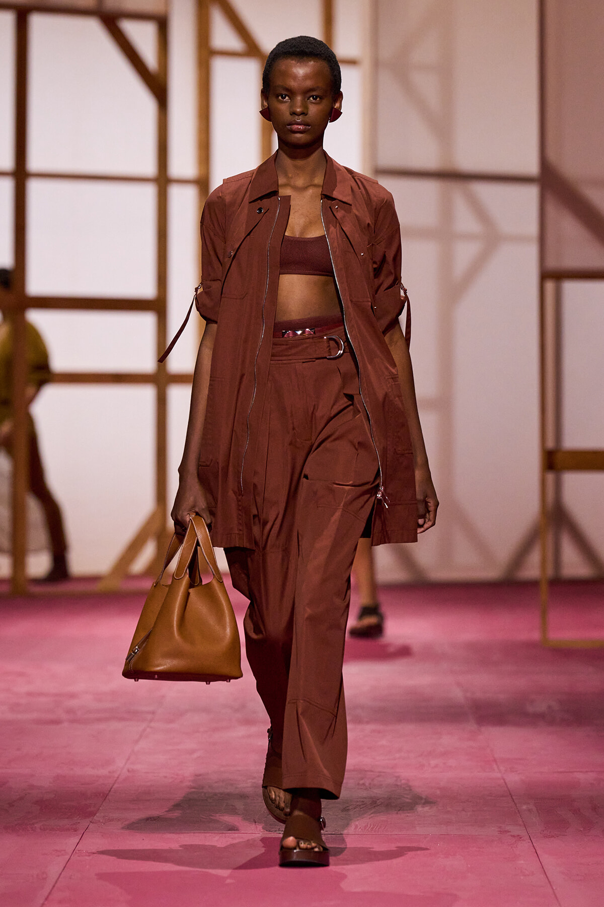 Model walks the runway in a rust-brown utility set: cropped top, oversized zip-front jacket, wide-leg pants with belt, and a tan handbag.