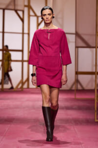 Model walks the pink runway in a magenta three-quarter-sleeve dress with a front buckle detail and black knee-high boots.