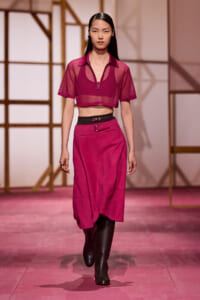 Model walking a fashion show wearing a magenta sheer crop top with matching midi skirt and black belt, black boots.