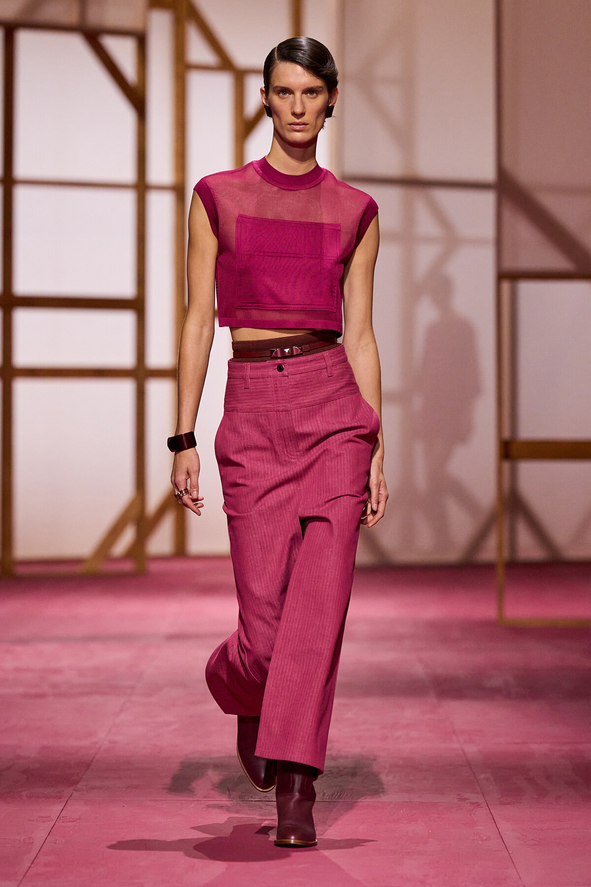 Model walking the runway in a magenta cropped sheer top with a matching high-waisted wide-leg pant set, burgundy boots, and a slim belt.