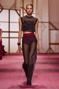 Model on a runway wearing a sheer black crop top and sheer burgundy pants with a pink belt and chunky bracelets.