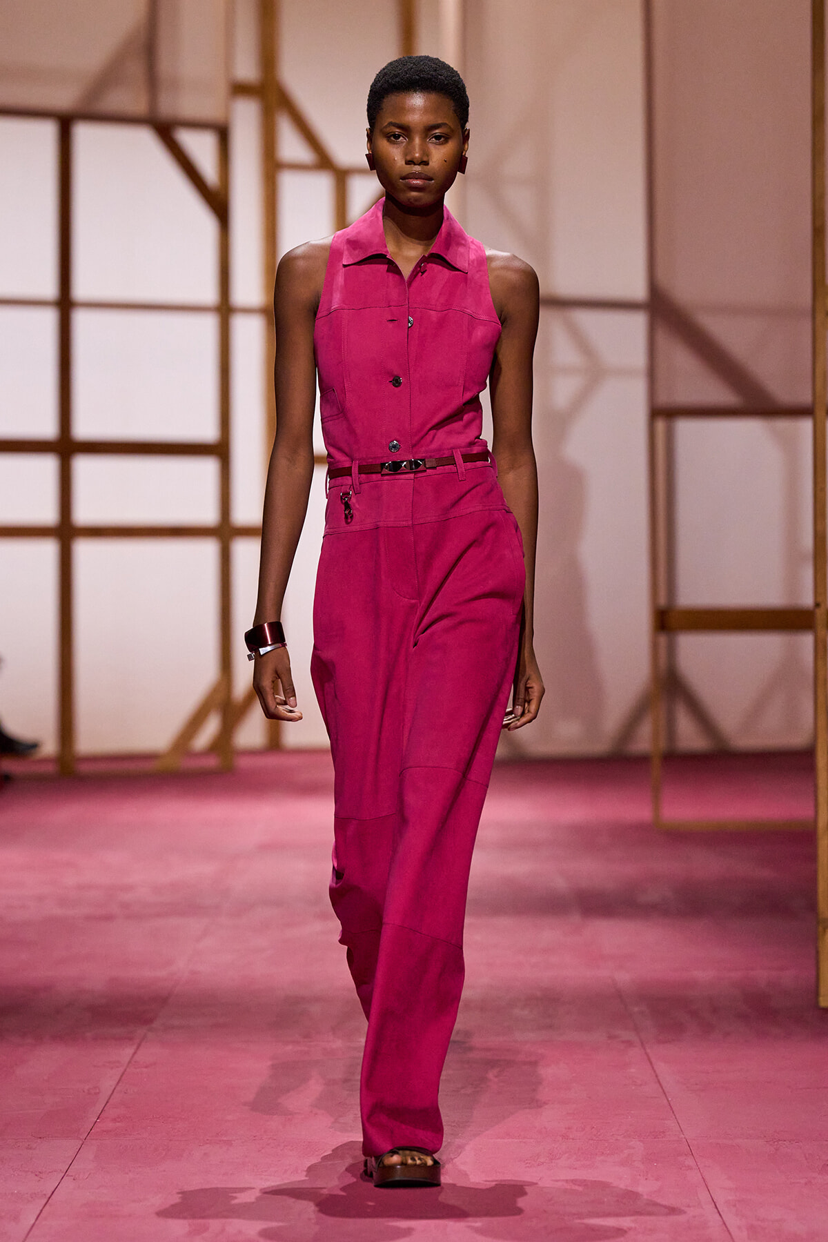 Model walking a runway in a sleeveless magenta jumpsuit with a black belt at the waist.