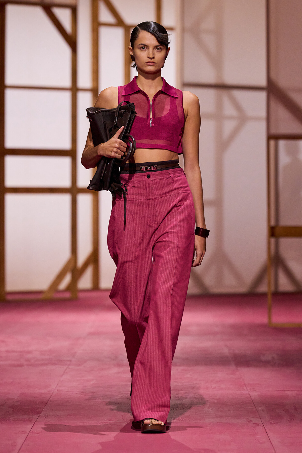 Model walks the runway in a pink cropped top and high-waisted wide-leg pants, carrying a black folded chair as a prop.