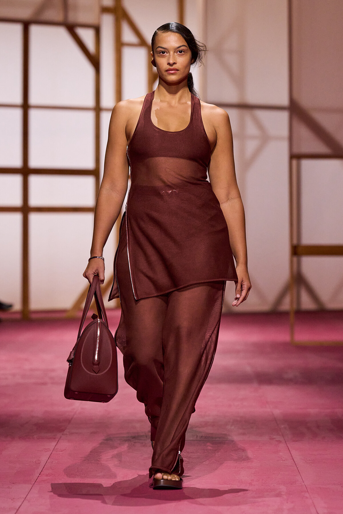 Model walking a runway in a burgundy sheer two-piece outfit with a matching burgundy handbag on a pink stage surface.