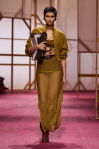 Model walks the runway in an olive-green coordinating outfit—a mustard bomber-style jacket over a cropped top and long skirt—while holding a beige and black handbag with rolled-up items and cables.