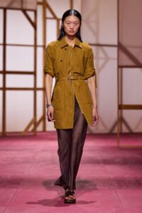 Model on a pink runway wearing a mustard belted trench coat with rolled sleeves and sheer black pants.