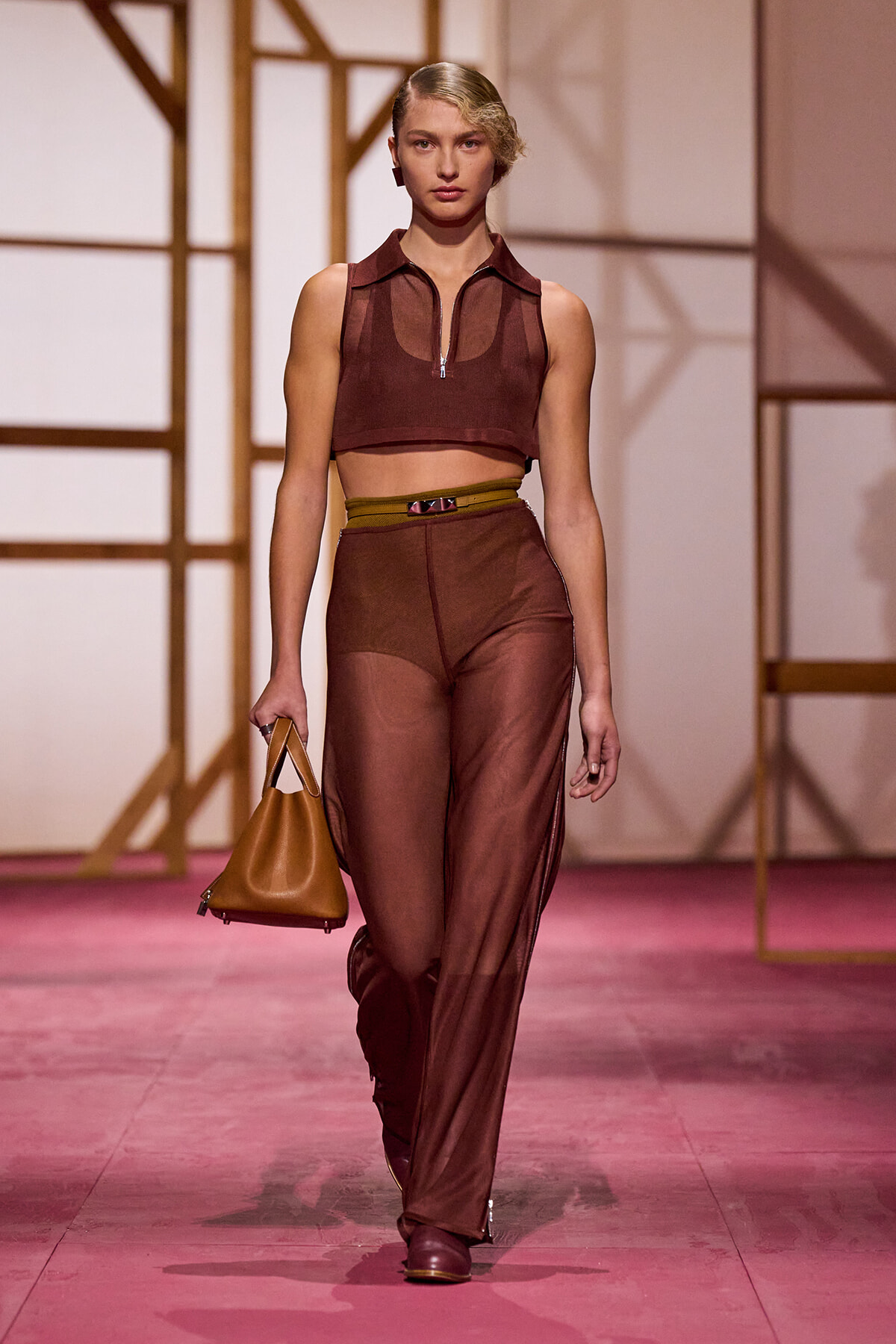 Model walks the runway in a brown two-piece set: cropped sleeveless zip-front top and sheer wide-leg pants, carrying a tan handbag.