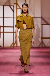 Model walking a fashion runway in a mustard cropped jacket, brown bralette, and high-waisted mustard pants with sandals.