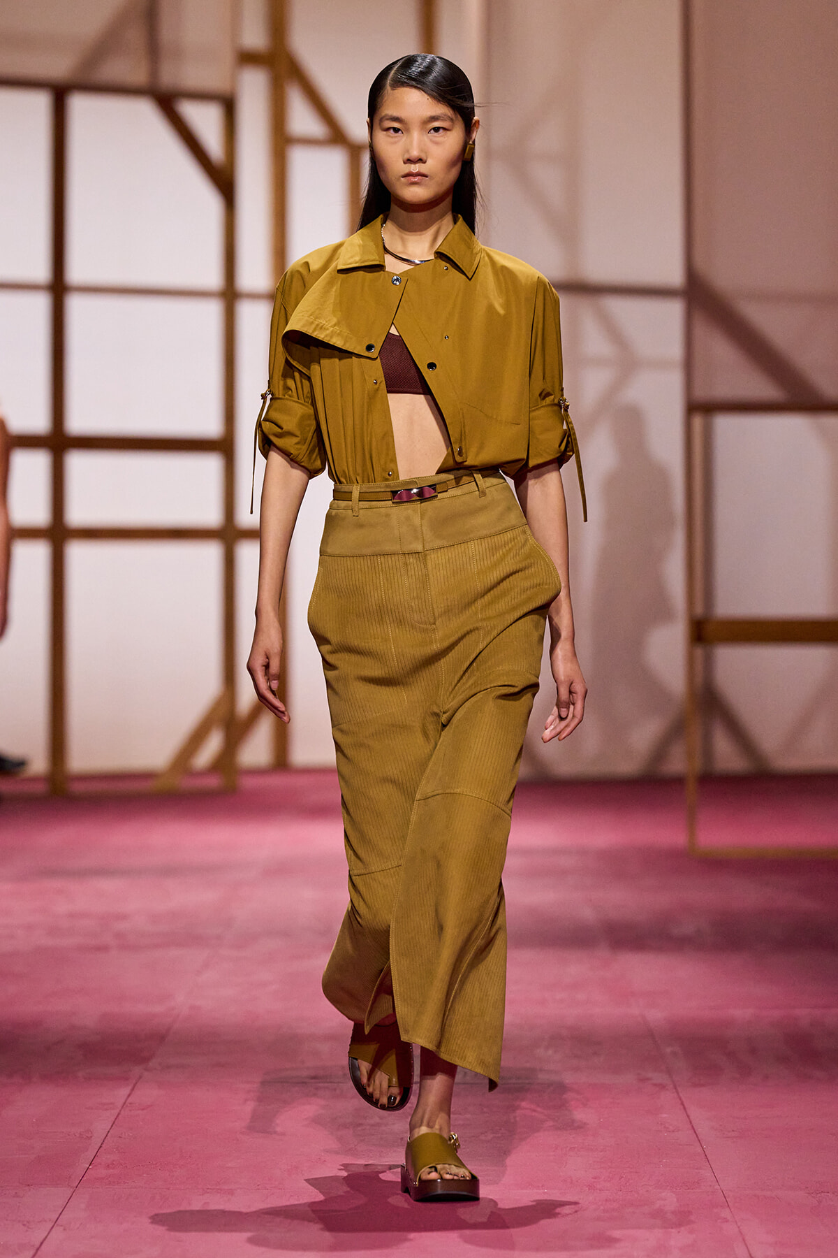 Model walking a fashion runway in a mustard cropped jacket, brown bralette, and high-waisted mustard pants with sandals.
