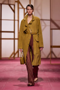 Model walking a fashion runway in a mustard trench coat with a belt, brown wide-leg pants, and a small mustard handbag.
