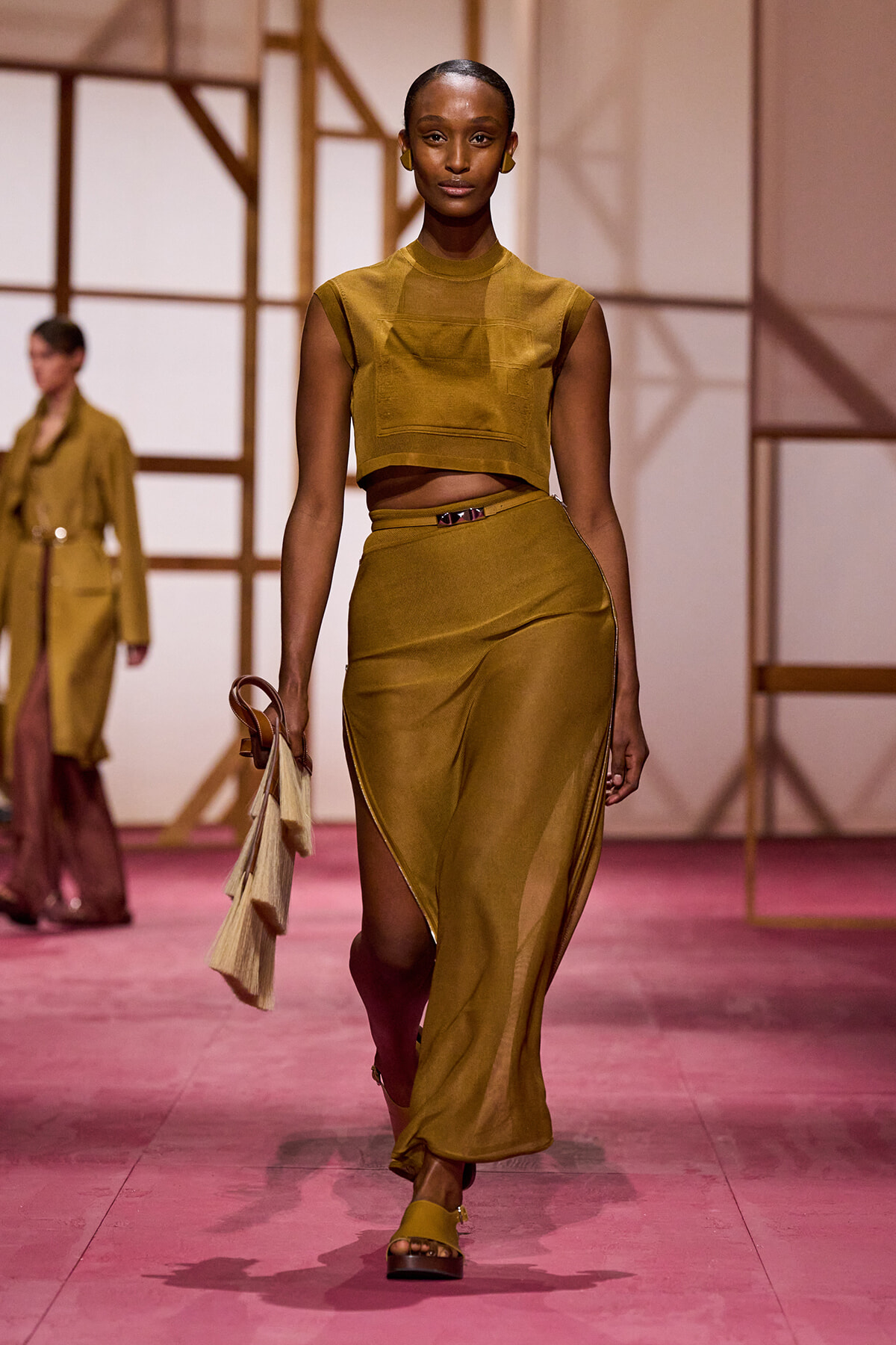 Model walking a fashion runway in a mustard crop top and matching high-waisted skirt with a front slit, carrying a beige fringe bag.