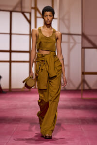 Model walks a fashion show wearing a mustard crop top and oversized cargo pants with a tied waist, carrying a round beige handbag on the left.