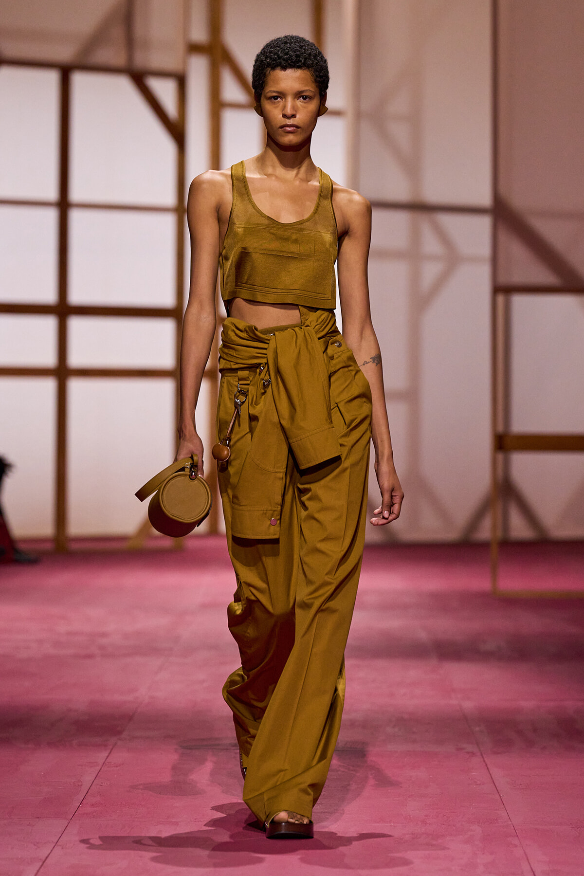 Model walks a fashion show wearing a mustard crop top and oversized cargo pants with a tied waist, carrying a round beige handbag on the left.