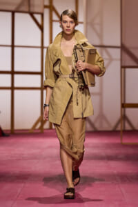 Model on a runway wearing a tan trench and beige outfit, carrying a structured tan bag under her arm.