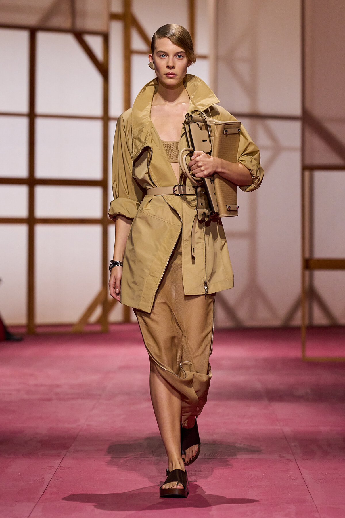 Model on a runway wearing a tan trench and beige outfit, carrying a structured tan bag under her arm.