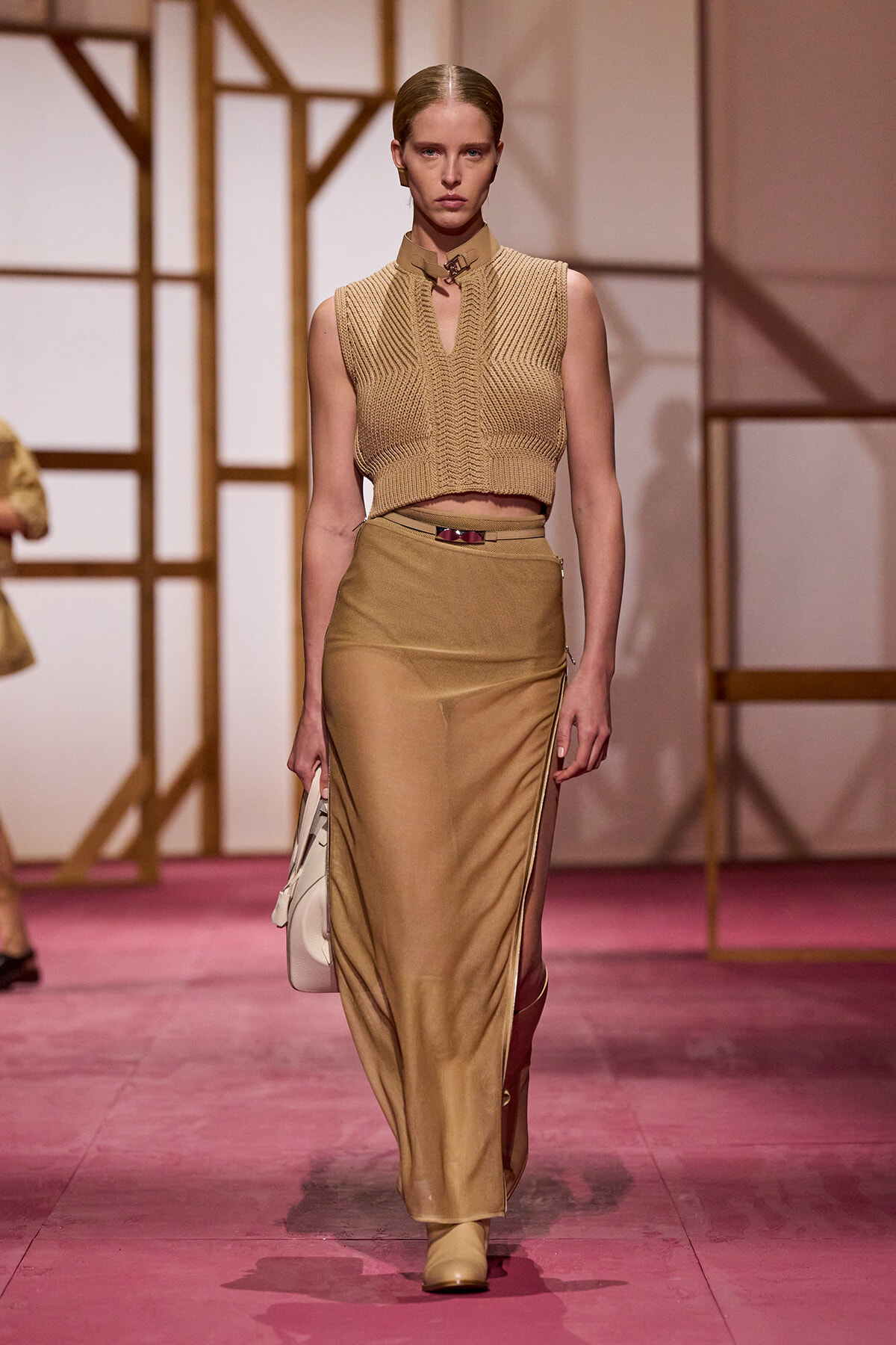 Model on a runway wearing a sleeveless beige knit crop top and a high-waisted tan satin skirt, carrying a light handbag.