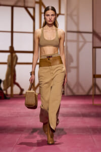 Model on a runway wearing a beige sports bra, high-waisted tan pants with a wide belt, tan boots, and a woven straw handbag.