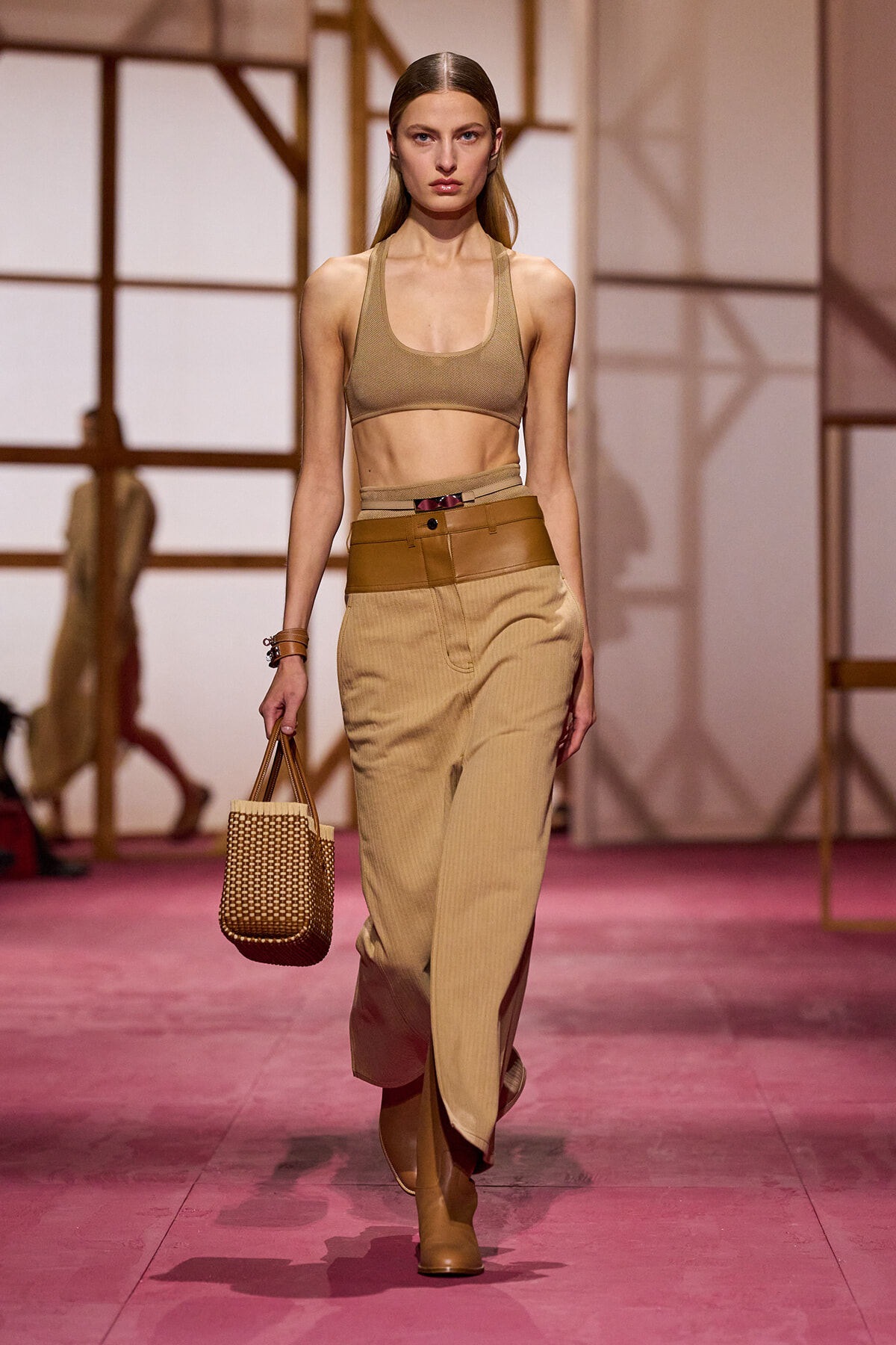 Model on a runway wearing a beige sports bra, high-waisted tan pants with a wide belt, tan boots, and a woven straw handbag.