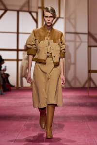 Model walking a fashion runway in a tan ensemble: cropped leather-trim jacket over a matching midi dress with knee-high boots on a pink catwalk.