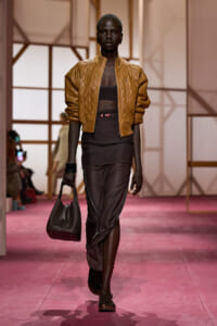 Model walks a runway wearing a tan quilted bomber jacket over a black crop top, paired with a dark draped skirt and a black handbag.