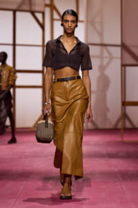 Model walking a fashion runway in a cropped sheer black top, tan leather pencil skirt with belt, and tan sandals, carrying a small woven handbag on a pink catwalk
