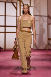 Model walks a fashion runway in a neutral tan crop top and matching high-waisted wide-leg pants, carrying a brown handbag.