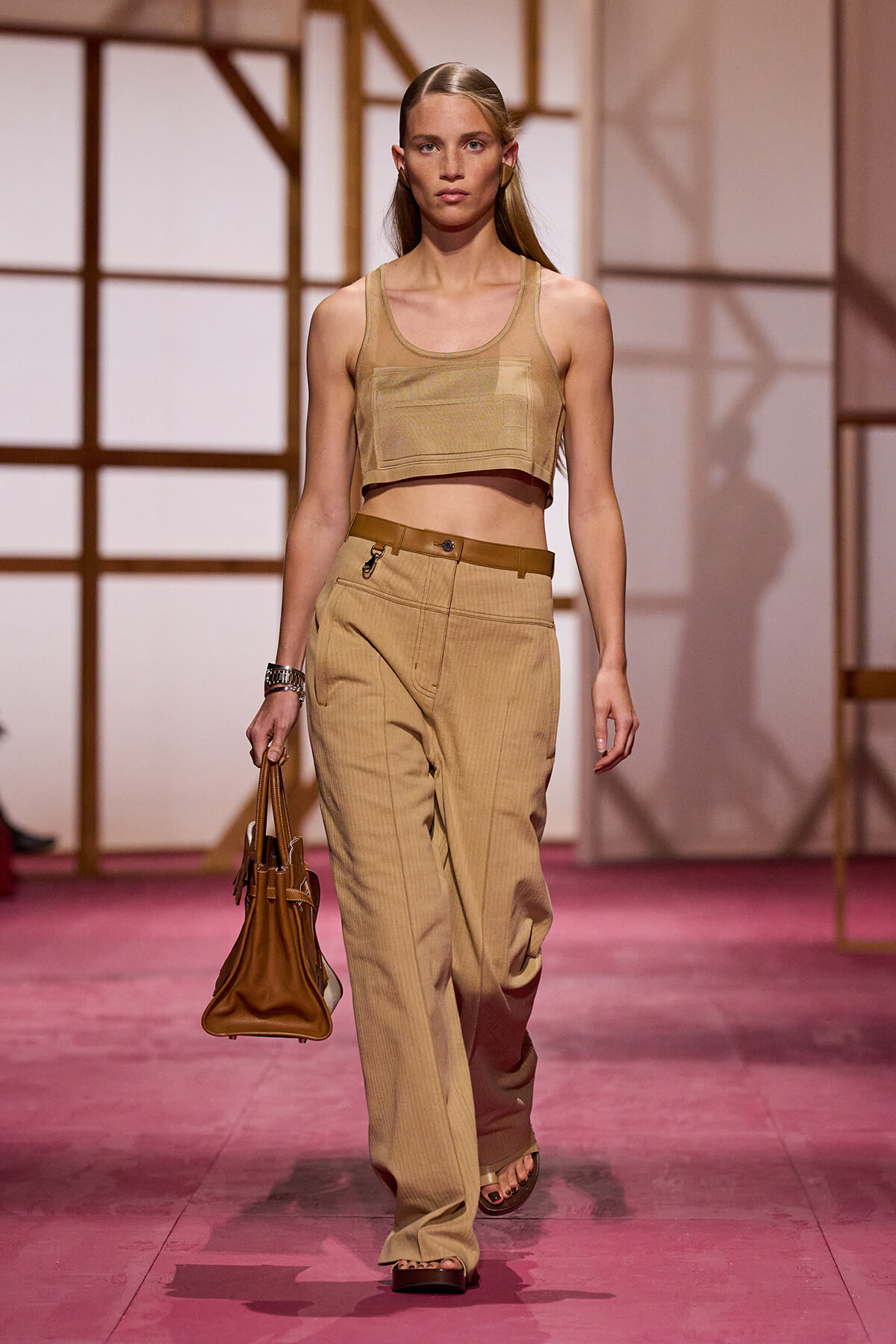 Model walks a fashion runway in a neutral tan crop top and matching high-waisted wide-leg pants, carrying a brown handbag.
