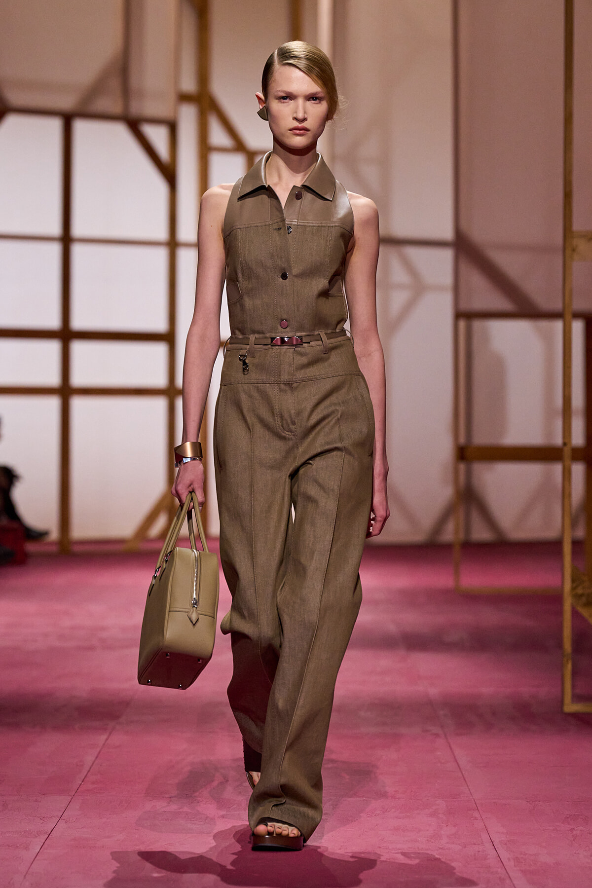 Model walks the runway in a sleeveless khaki utility jumpsuit with a matching belt, carrying a beige handbag.