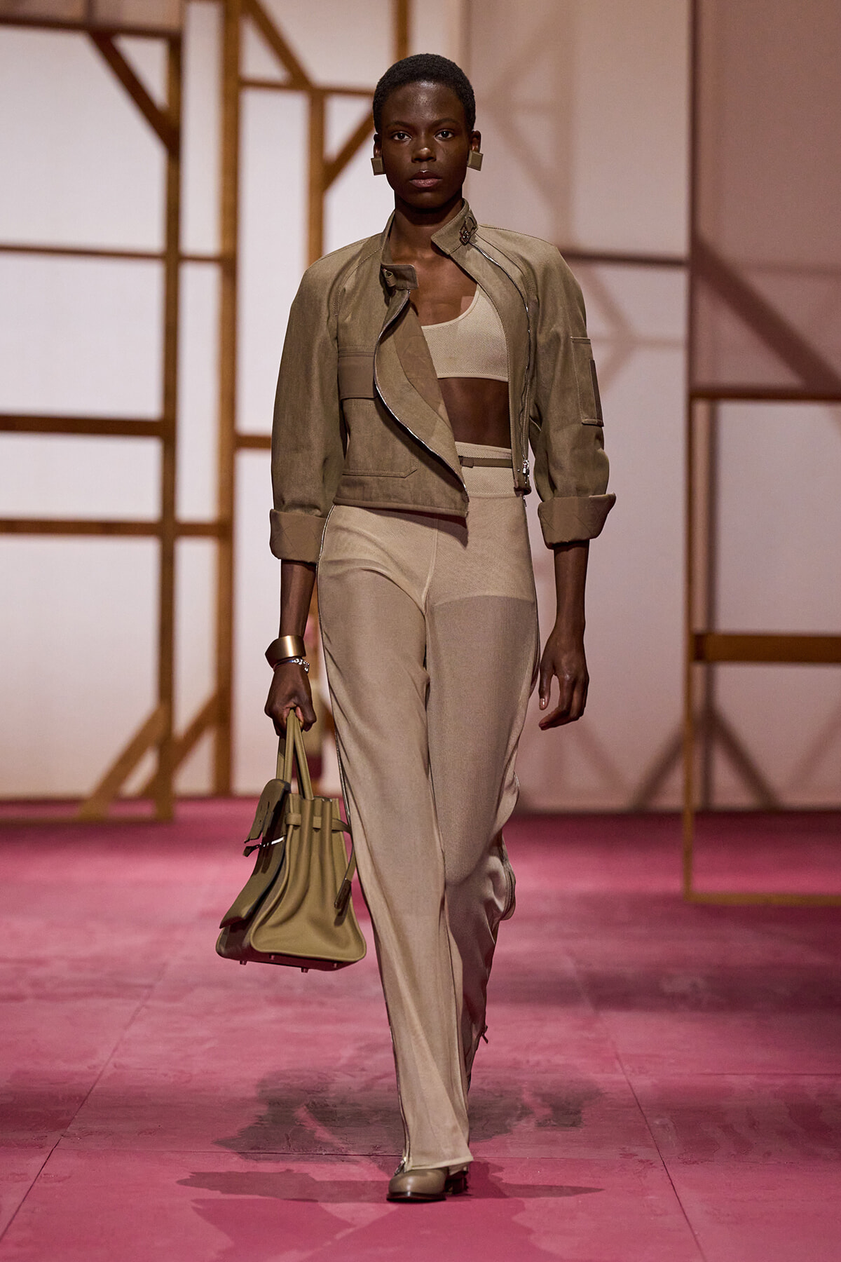 Model on a fashion runway wearing a tan cropped jacket, beige crop top, high-waisted beige pants, and a tan handbag.