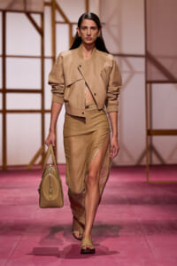Models wears beige cropped jacket with matching high-split skirt and tan accessories on a runway, carrying a beige bag.