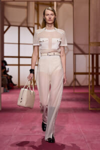 Model walking a fashion runway in a sheer cream top with pockets and high-waisted wide-leg pants, carrying a beige handbag.