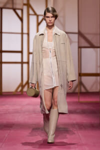 Model walking a pink runway in a beige oversized coat over a sheer cream ensemble, carrying a small round tan handbag and wearing knee-high boots.