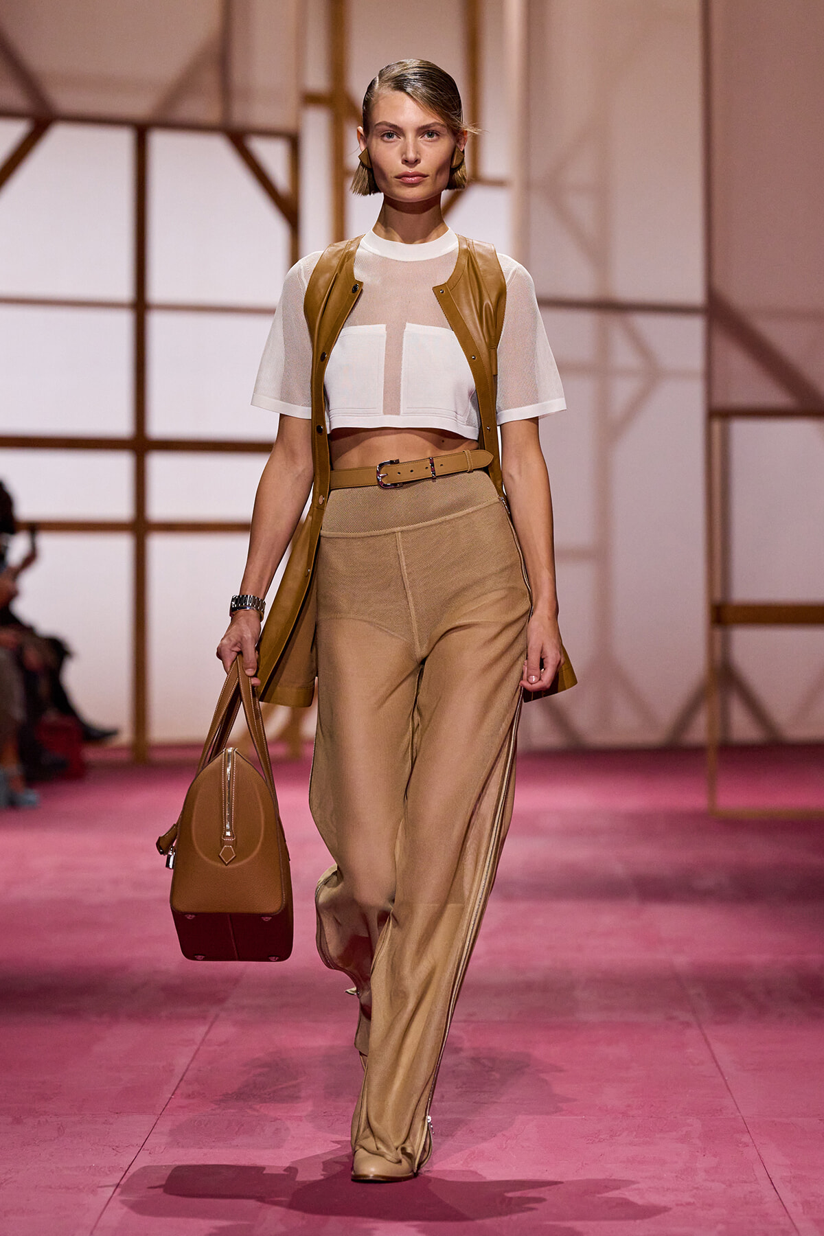 Model walking the runway in a tan leather sleeveless vest over a sheer white crop top, paired with wide tan pants and a tan belt, carrying a tan handbag on a pink catwalk.