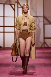 Model walks the runway in a beige trench coat over a brown bra top and high-waisted briefs, brown knee-high boots, and a tan handbag.
