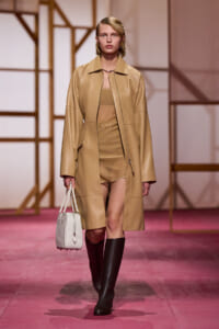 Model walks the runway in a tan leather coat over a beige dress, paired with black knee-high boots and a white handbag.