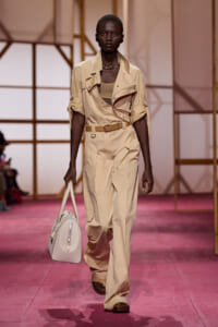 Model walking the runway in a beige utility jumpsuit with rolled sleeves, carrying a white tote bag.