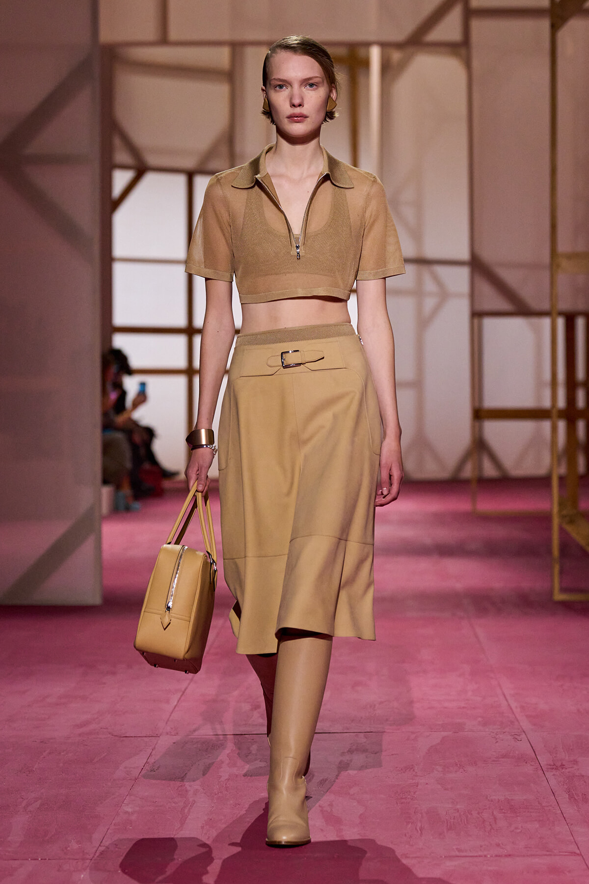 Model walking a runway in a tan cropped top, matching high-waist skirt, knee-high boots, and a beige handbag.