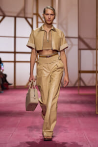 Model walks a fashion runway in a beige cropped jacket and matching high-waisted pants, carrying a beige handbag.