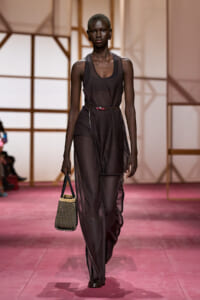 Female model walking a fashion runway in a sheer black jumpsuit with a pink belt, carrying a woven handbag on a pink catwalk.