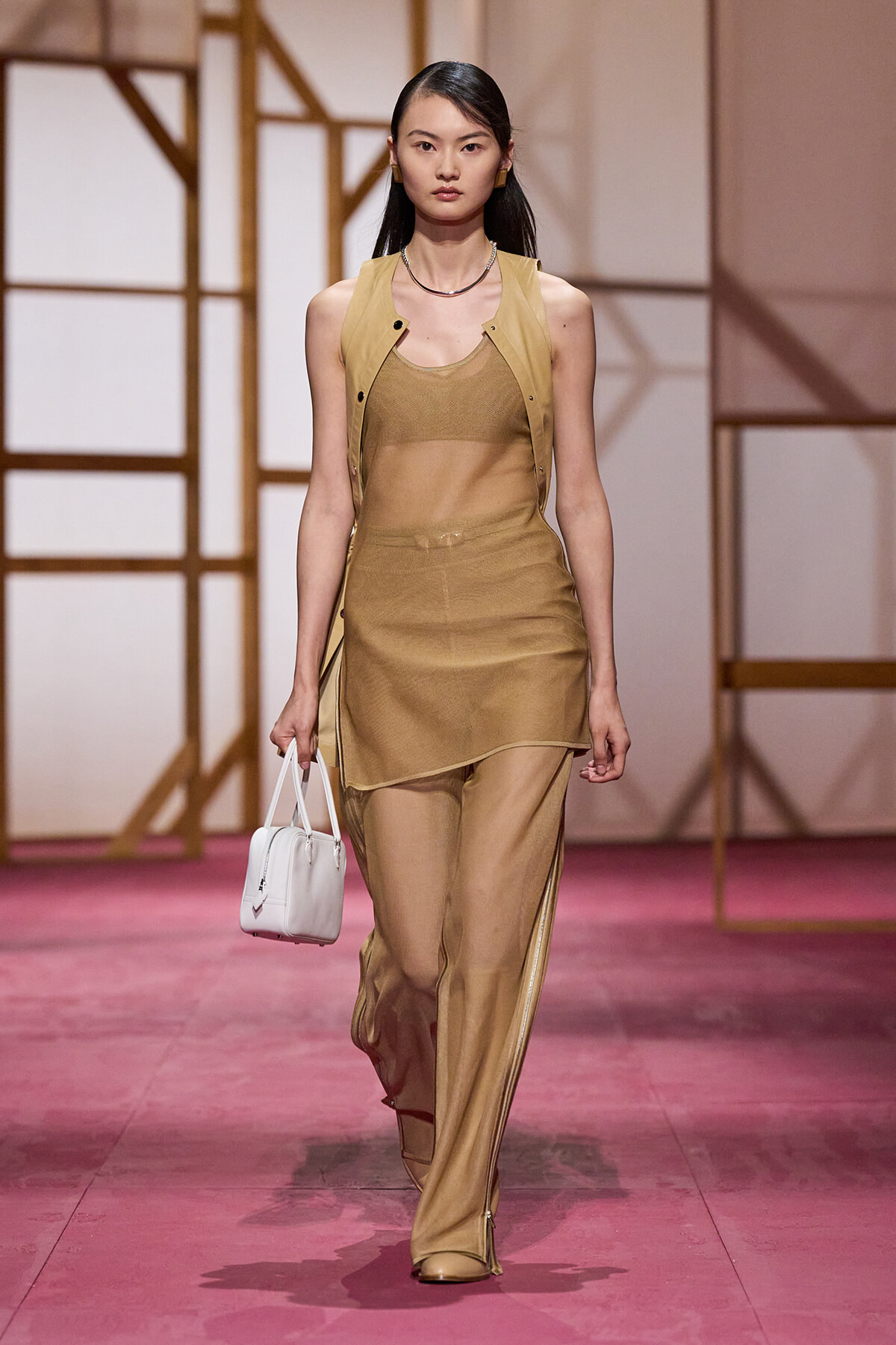 Model walking a runway in a tan mesh set: sleeveless vest over a sheer top and matching layered skirt with wide-leg pants, carrying a white handbag