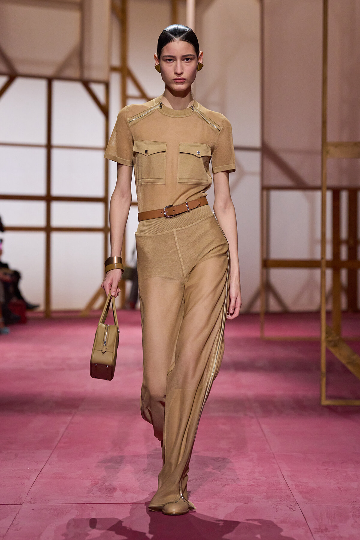 Model walks the runway in a camel-toned utility outfit: short-sleeve top with front pockets, matching wide-leg pants, and a brown belt, carrying a small beige bag.