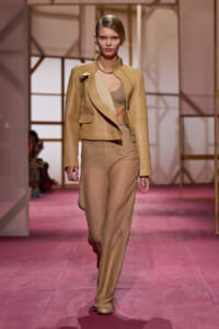 Model walking the runway in a tan leather blazer over a beige crop top and matching wide-leg pants on a pink catwalk.