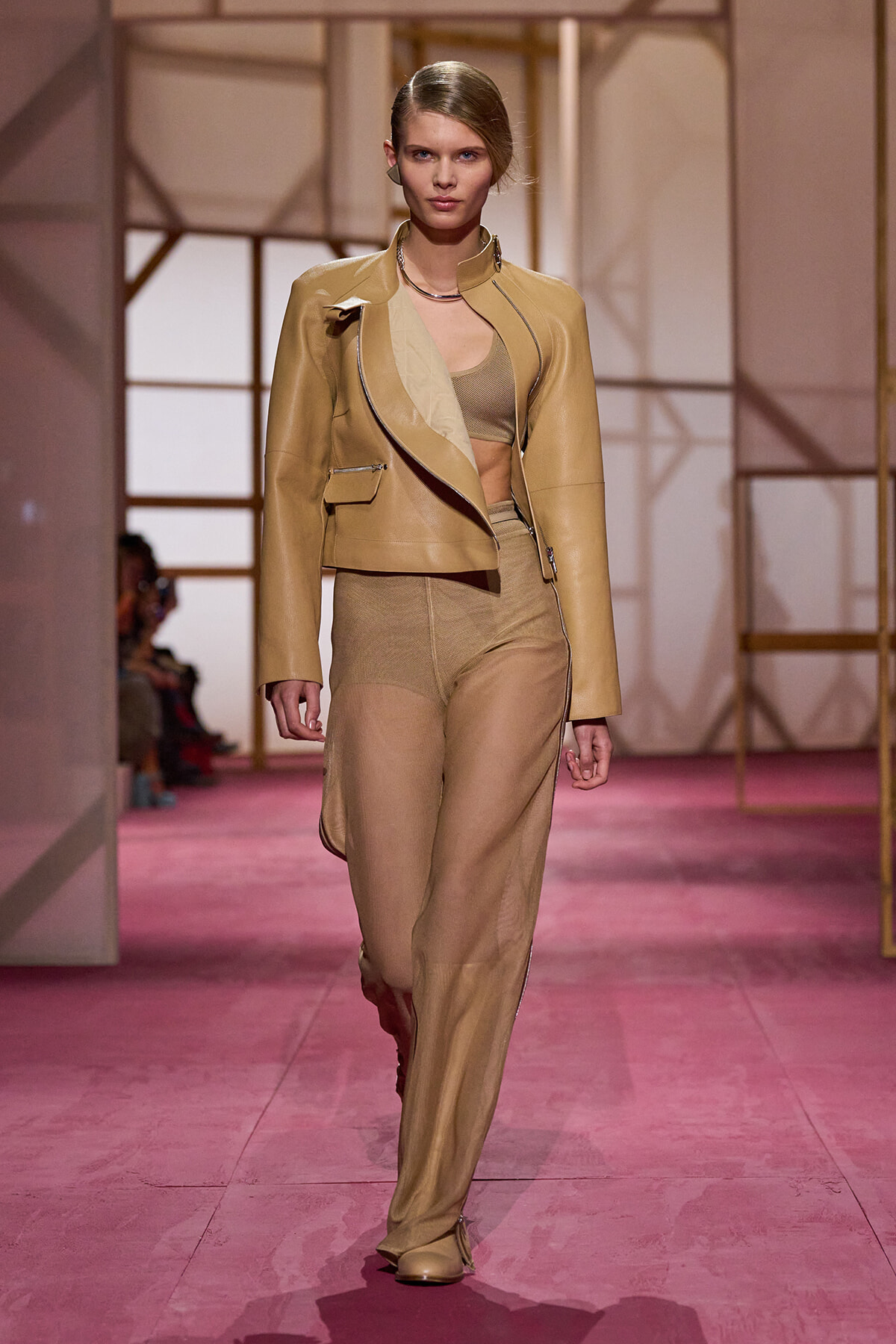 Model walking the runway in a tan leather blazer over a beige crop top and matching wide-leg pants on a pink catwalk.