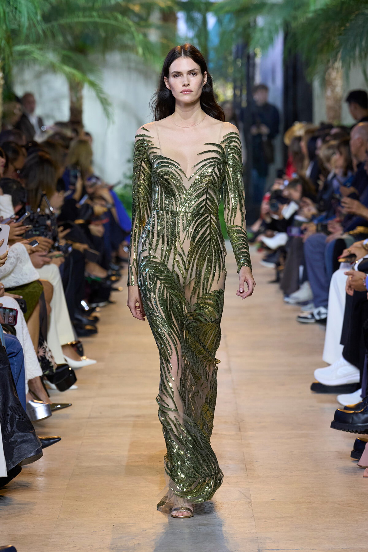 Model walking runway in a sheer green sequin gown with palm leaf embroidery, crowd on both sides.