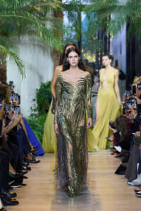 Model walks a green sequined gown with palm leaf pattern down a runway lined by photographers and palm decor.