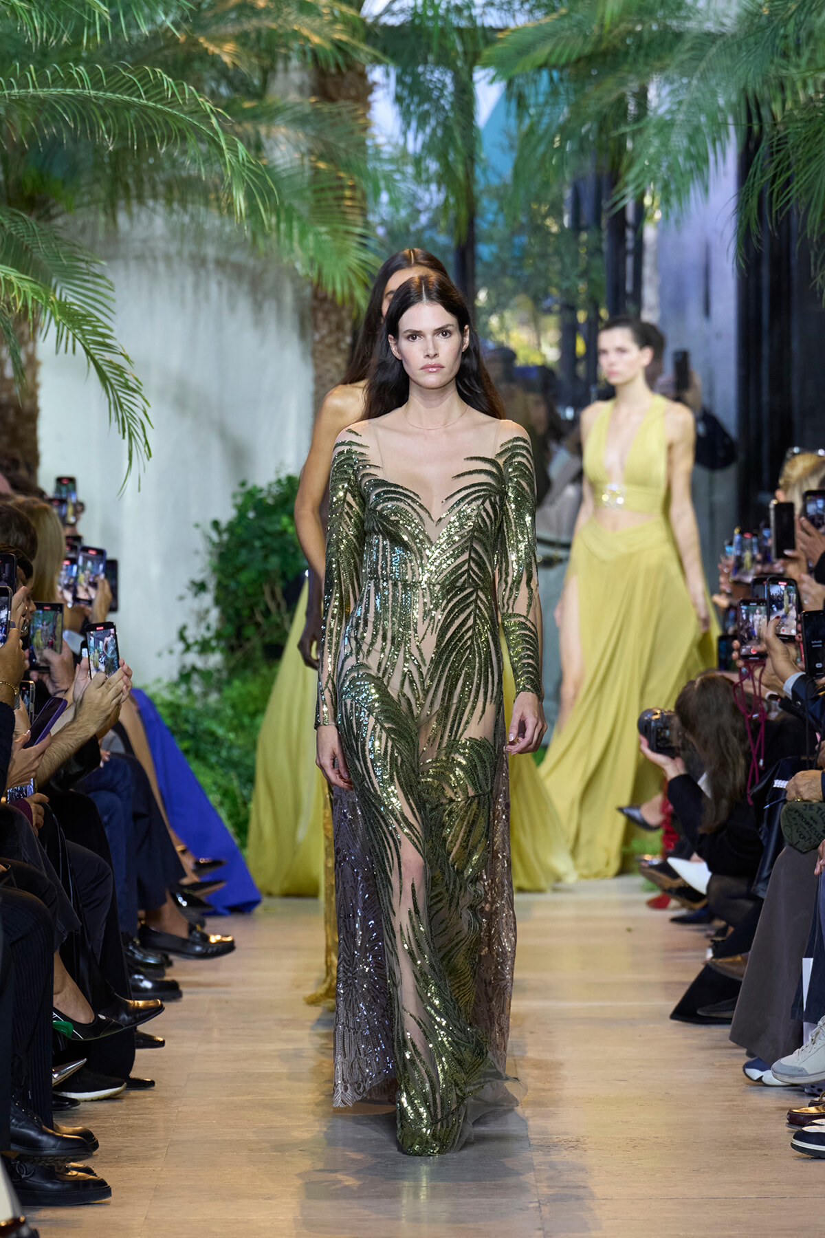 Model walks a green sequined gown with palm leaf pattern down a runway lined by photographers and palm decor.