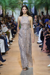 Model walks a silver beaded halter gown down a runway at a fashion show with spectators on both sides of the aisle.