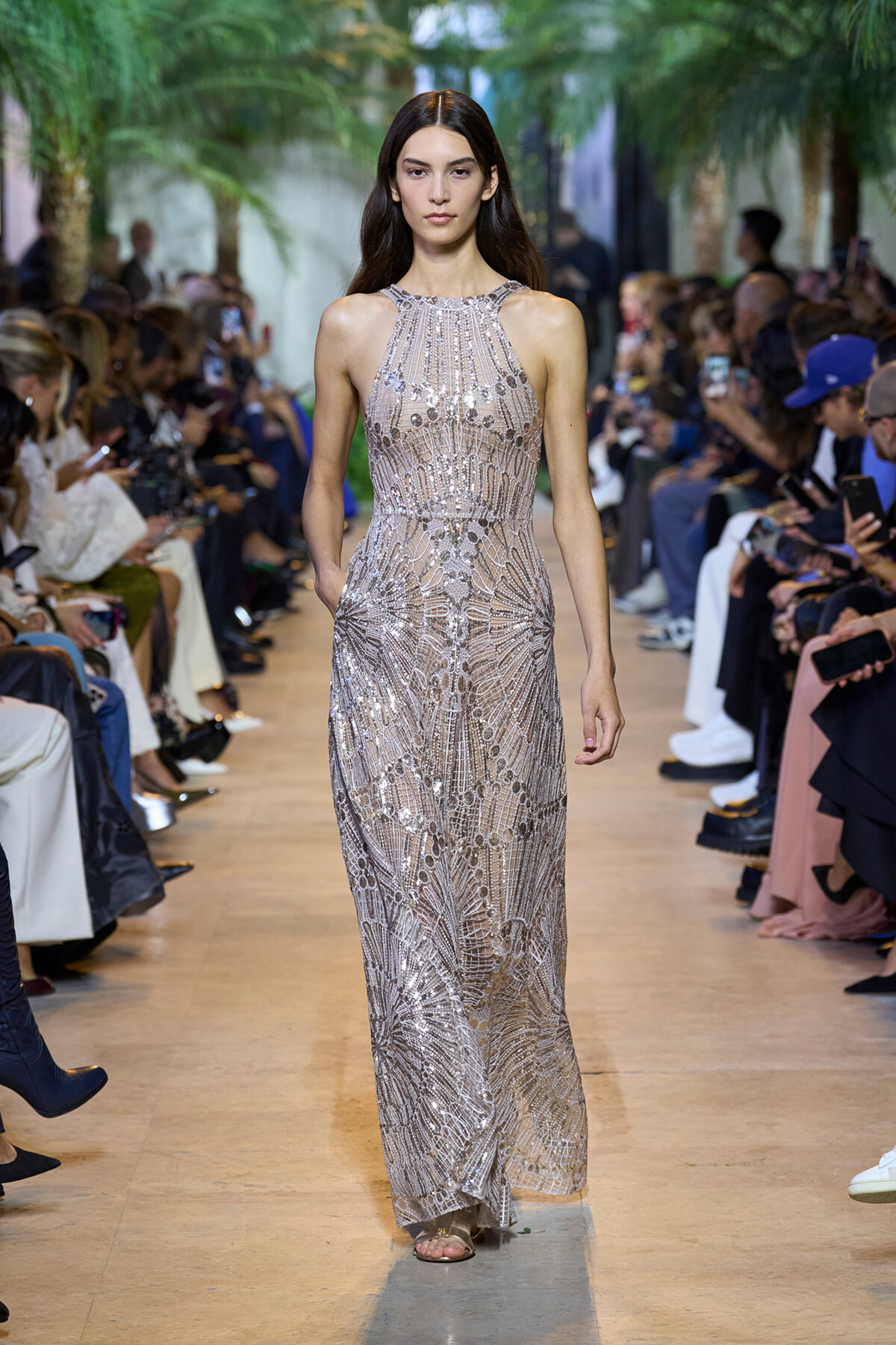 Model walks a silver beaded halter gown down a runway at a fashion show with spectators on both sides of the aisle.